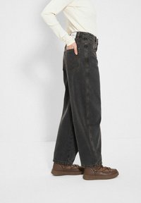 Szerokie, ciemnoszare dżinsy z wysokim stanem, z pomarańczowymi szwami i tylnymi kieszeniami, zestawione z brązowymi sznurowanymi butami.