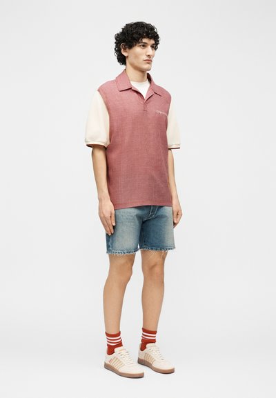 Jeune homme portant un polo sans manches à carreaux rouges, une chemise à manches courtes crème, un short en denim, des chaussettes rayées rouges et des baskets crème, debout devant un fond blanc.