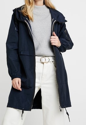 Kobieta w granatowej kurtce z kapturem na szarym swetrze, białych spodniach z białym paskiem z podwójną klamrą, stoi z jedną ręką na kurtce.