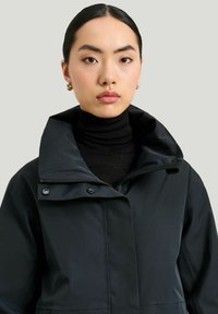 Svart vattentålig jacka med hög krage, dragkedja och tryckknappar. Har en slät textur och en figurnära design.