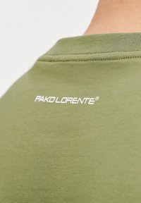 Oliwkowa koszulka z miękkiego materiału, z okrągłym dekoltem i białym logo "PAKO LORENTE" na plecach. Gładka tekstura i minimalistyczny design.