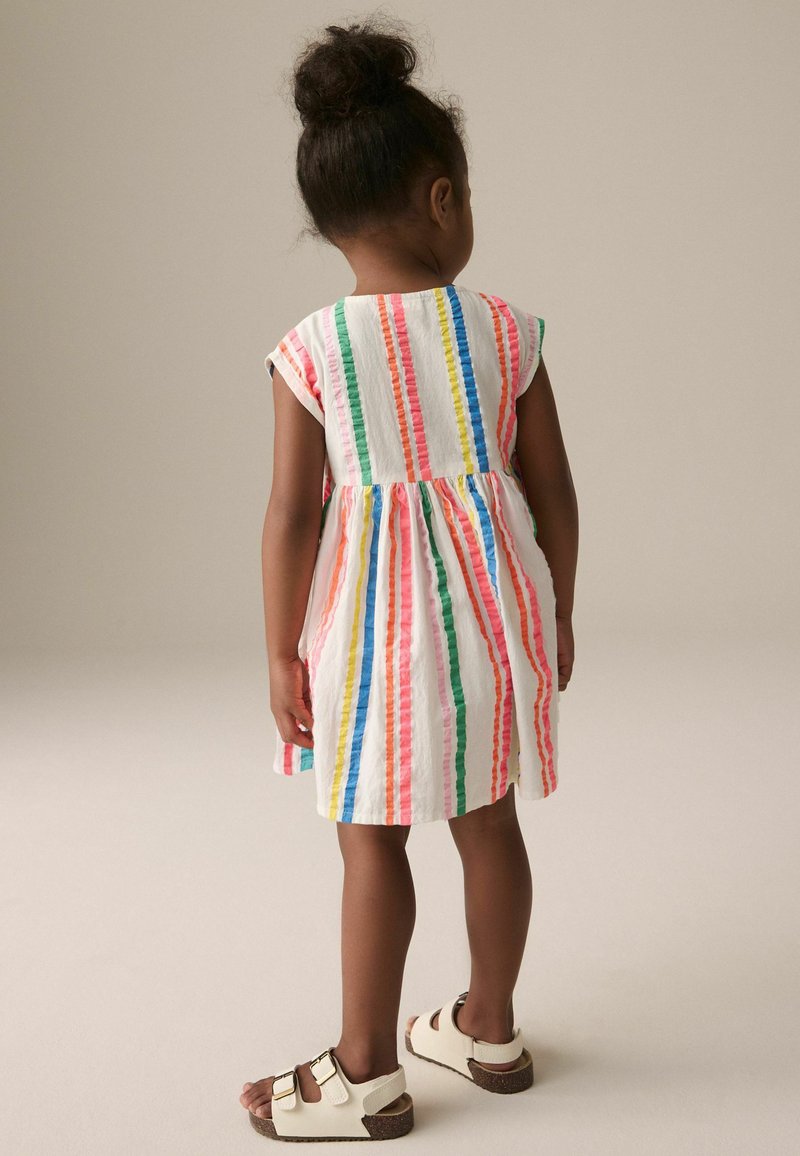 Robe rayée colorée à manches courtes, en tissu léger. Les rayures sont de couleur rose, bleue, verte et orange. Portée avec des sandales blanches.