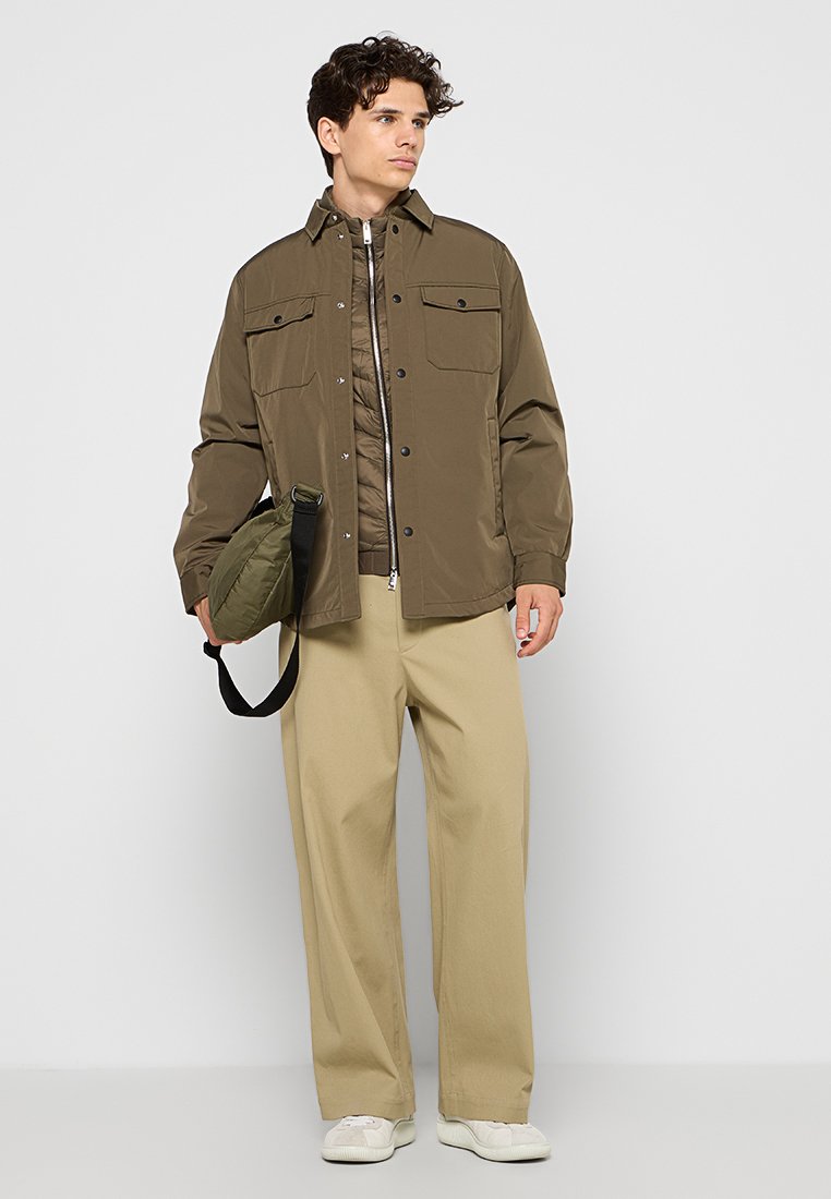 Veste vert olive avec deux poches poitrine, fermeture à boutons et une coupe légèrement oversize. Pantalons beiges, baskets blanches et sac à épaule vert.