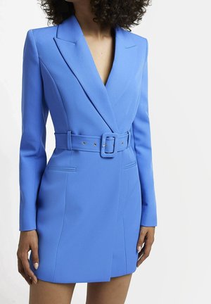 Mujer con un vestido-blazer azul ajustado de manga larga, con cinturón y escote en V pronunciado, de pie sobre un fondo blanco liso.