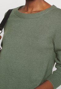 Grön stickad tröja med rund halsringning och ribbade ärmslut. Mjuk textur med en liten lyster och avslappnad passform. Svart rem synlig.