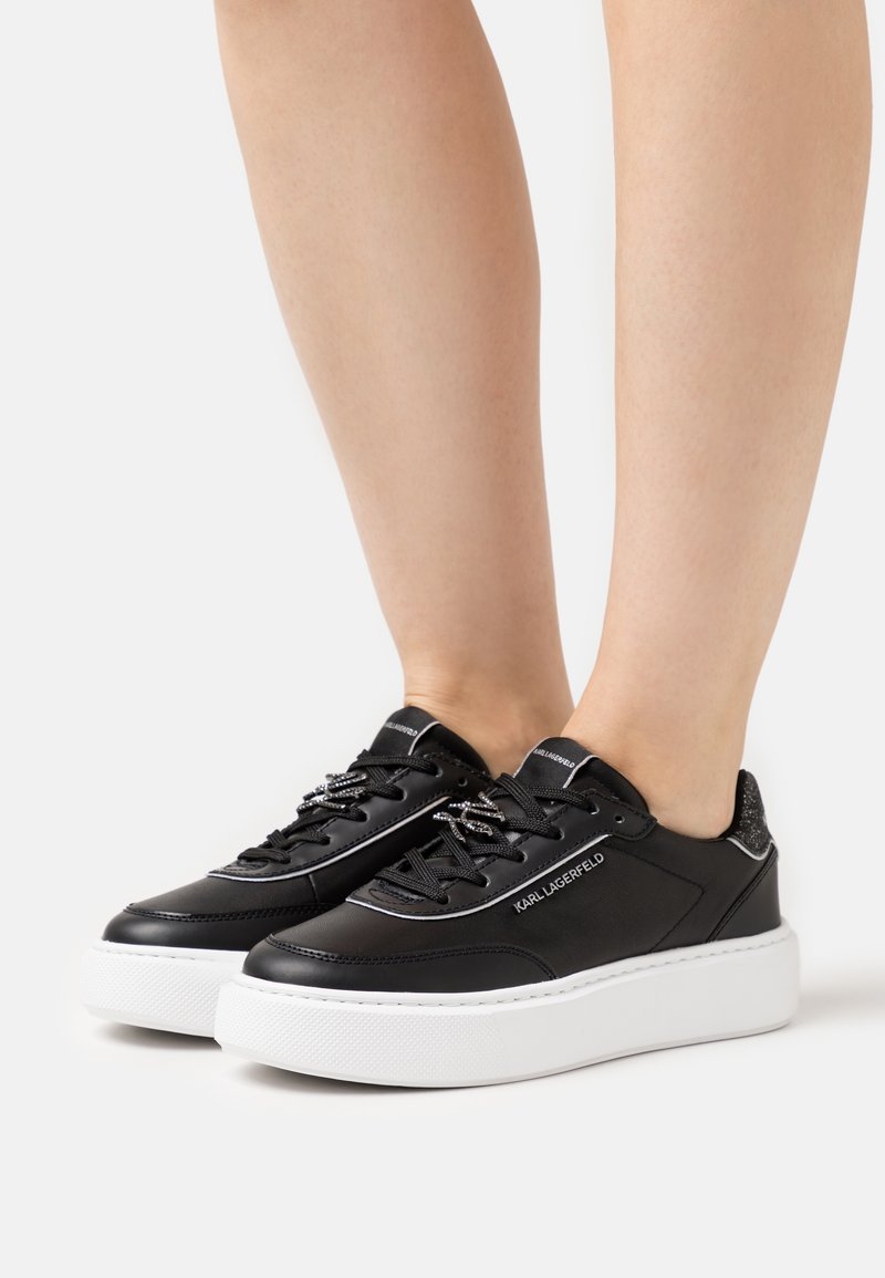 Mustat nahkalenkkarit, joissa on valkoinen kumipohja, metallinhohtoiset nauhat ja sivussa hillitty logo, sekä kimaltelevat yksityiskohdat kannassa.