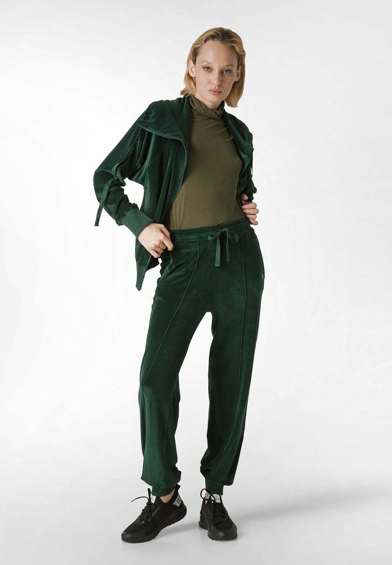 Tuta in velluto verde con giacca con zip e pantaloni con cordino. La parte superiore presenta un colletto alto e i pantaloni hanno cuciture laterali. Sneakers nere.