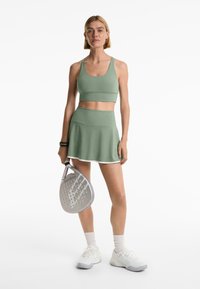 A zöld sportmelltartó és szoknya szett sima anyaggal, testhez simuló kialakítással és fehér díszítéssel rendelkezik. A modell egy szürke pingpong ütőt tart.