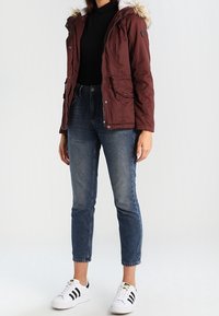 Bordowa ocieplana kurtka z odpinanym kapturem z sztucznego futra, zestawiona z ciemnoniebieskimi jeansami i białymi trampkami z czarnymi paskami.