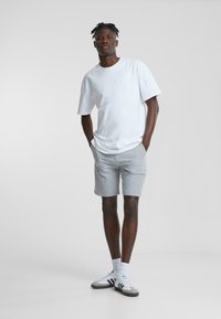 Camiseta blanca de manga corta, pantalones cortos a cuadros grises y zapatillas grises con detalles en negro. El modelo está de pie con las manos en los bolsillos. Textura suave.
