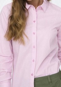 Camicia a righe rosa chiaro con bottone frontale, colletto a punta e maniche lunghe. I bottoni sono di una tonalità di rosa abbinata.