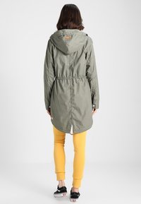 Oliwkowa kurtka z kapturem, z wcięciem w talii, długimi rękawami i lekko wydłużonym tyłem. Posiada zatrzaski oraz logo.