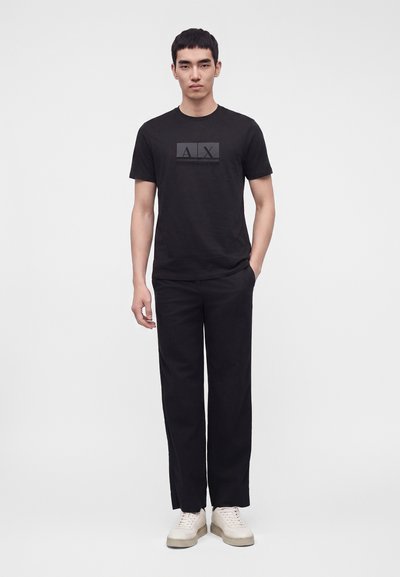 Млад мъж, облечен с черна тениска Armani Exchange, черен панталон и бели маратонки, стои срещу светъл фон.