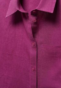 Chemise en lin magenta avec col pointu, fermeture à boutons et poche poitrine. Le tissu présente une surface légèrement texturée et un aspect doux.