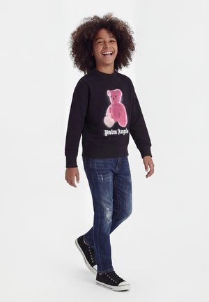 Copil zâmbitor cu păr creț care merge, purtând un hanorac negru cu grafică urs roz și textul „Palm Angels”, blugi albaștri și adidași negri.