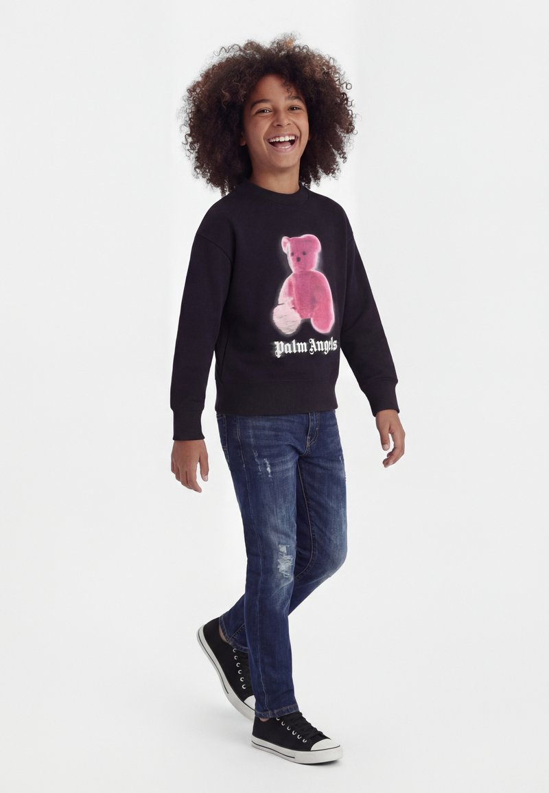 Παιδί που χαμογελά με σγουρά μαλλιά περπατά, φοράει μαύρο φούτερ με ροζ γραφικό αρκούδας και κείμενο «Palm Angels», μπλε τζιν και μαύρα αθλητικά παπούτσια.