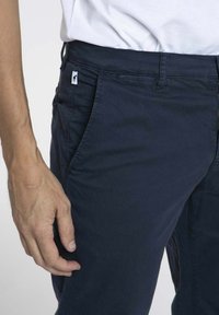 Chinos en coton bleu marine avec poches latérales, une texture lisse et une petite étiquette logo sur la taille, équipés d'une passante standard pour la ceinture.