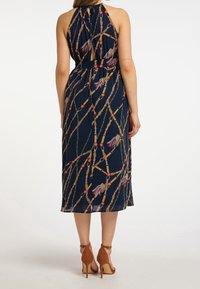 Marinblå halterneckklänning med en urklippt rygg, som har ett mönster i guld och röd kedja. Tyget är lätt med en mjuk textur. Bruna klackar med ankelrem.