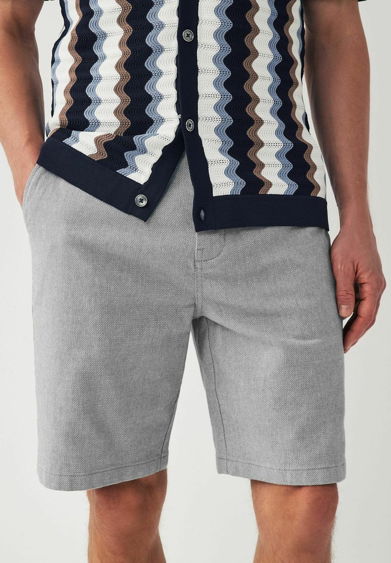 Chemise à manches courtes avec un motif en zigzag bleu, marron et blanc ; associée à un short gris clair en tissu texturé.