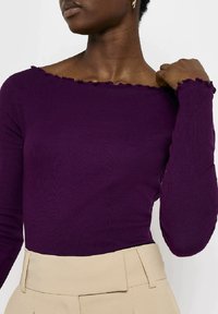 Fioletowa, żeberkowana bluzka z długim rękawem, z falbaną na dekolcie typu off-shoulder i dopasowanym krojem, w połączeniu z kremowymi spodniami z wysokim stanem.