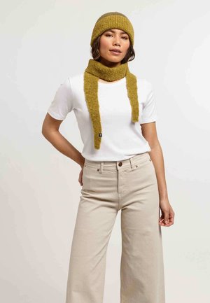 Woman wearing a green knitted hat and scarf, white short-sleeve shirt, and beige high-waisted wide-leg pants, standing with one hand on hip.