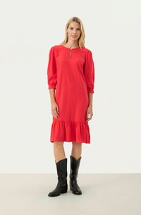 Röd knälång klänning med puffärmar, nyckelhålsurklipp och volangkant. Bärs med svarta knähöga stövlar, med slät textur och livfull färg.