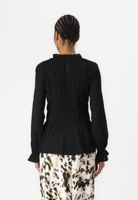 Black long-sleeve blouse with smocked texture, ruffled collar, keyhole back, and fitted waist. Paired with a patterned, flowing skirt.