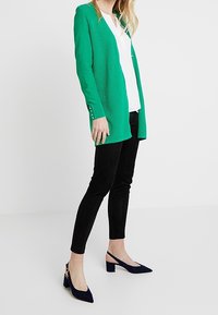 Zelený strukturovaný cardigan s výstřihem do V, kombinovaný s bílou blůzou a černými slim-fit kalhotami. Obuv zahrnuje modré slingback podpatky.