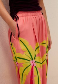 Pantalon rose avec un imprimé palmier en vert et jaune. Il dispose d'une taille élastique et de poches latérales, fabriqué en tissu léger.