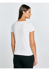 Biała bawełniana koszulka z krótkimi rękawami i zaokrąglonym dekoltem, dopasowany krój i gładka faktura. Noszona z czarnymi spodniami, widoczna od tyłu.