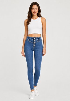 Kvinde iført en hvid kort top, højtskårne blå skinny jeans med flere knapper og hvide sneakers, stående mod en ensfarvet baggrund.
