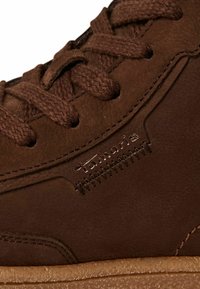 Zapatilla de cuero marrón con acabado texturizado, que presenta cordones marrones, detalles de costura y un logotipo de la marca visible grabado en el lateral.