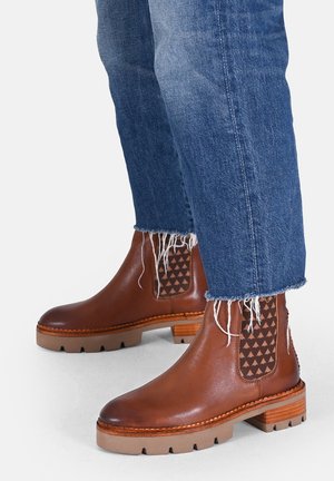 Stivali alla caviglia in pelle marrone con un pannello elastico testurizzato a motivo triangolare e una suola in gomma robusta di colore chiaro. Jeans con orli sfilacciati.