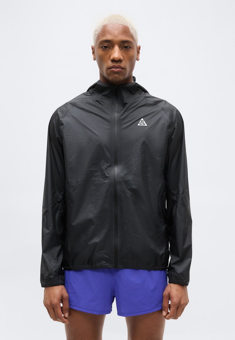 Νεαρό άτομο με κοντά ξανθά μαλλιά φοράει μαύρο ελαφρύ μπουφάν με το λογότυπο ACG και φωτεινά μπλε αθλητικά σορτς σε απλό φόντο.