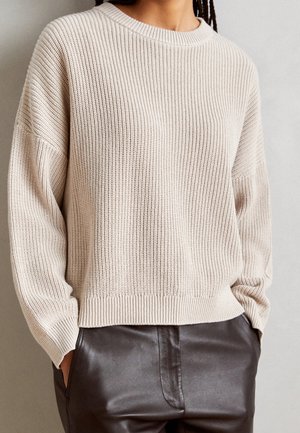Personne portant un pull en maille côtelée beige et un pantalon en cuir marron foncé avec les mains dans les poches, sur un fond neutre.