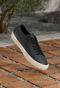 Black leather sneaker with round toe and flat sole, featuring black laces, a smooth texture, and a contrasting cream-colored rubber sole.