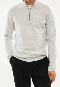 Homme portant un sweat-shirt gris clair à demi-fermeture éclair et un pantalon noir, debout avec une main dans la poche et l'autre main détendue le long du corps.