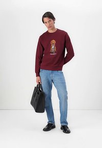 Sweatshirt cor de vinho com gráfico de cão e texto "HACKETT", combinado com jeans azuis e sapatos de couro pretos, segurando uma mala tote preta.