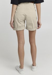 Shorts beige à revers avec une texture lisse, dotés de poches arrière et d'une taille haute, associés à des baskets blanches sur un fond uni.