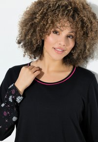 Černý top s dlouhými rukávy, který má růžový lem kolem výstřihu a na rukávech barevné vzory sněhových vloček pro zajímavý detail.