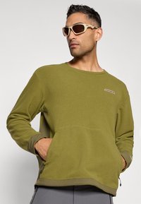 Camicia a maniche lunghe di un verde oliva testurizzato con scollo rotondo, tasca a busta sul davanti e rifiniture a contrasto di un verde scuro.