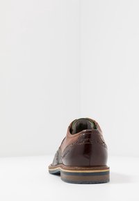 Chaussure en cuir marron avec des accents texturés gris, un bout arrondi, des coutures détaillées et une semelle superposée aux nuances de bleu et de marron.