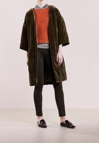 Olivgrüner Samtmantel mit weit geschnittenen Ärmeln, orangefarbener strukturierter Pullover, karierten schwarzen Hosen und glänzenden schwarzen Loafers mit Hardware-Details.