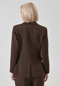 Blazer marrone a righe sottili con design aderente, spacco singolo sul retro e maniche lunghe. Tessuto dalla texture liscia, silhouette sartoriale, dettagli minimali.