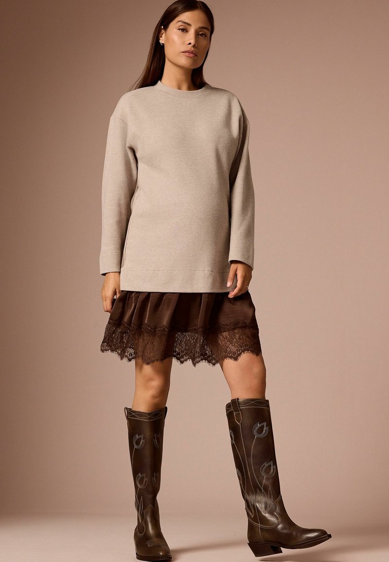 Vrouw draagt een beige top met lange mouwen, een bruine rok met kanten afwerking en donkerbruine geborduurde kniehoge laarzen op een beige achtergrond.