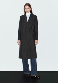 Cappotto grigio doppiopetto in tessuto texturizzato, dal taglio lungo fino al ginocchio, con due tasche frontali, abbinato a jeans a gamba larga e sandali neri.