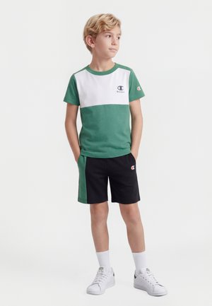 Garçon blond debout les mains dans les poches, portant un t-shirt vert et blanc à manches courtes, un short noir et vert, des chaussettes blanches et des baskets blanches.