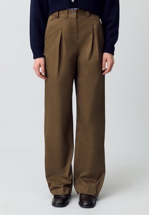 Brune højtaljede bukser med vide ben og folder, båret med sorte sko og en mørk marineblå bluse med lange ærmer, stående foran en ensfarvet baggrund.
