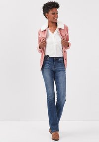 Chaqueta de pana rosa con cuello de piel sintética blanca, llevada sobre una camisa blanca, combinada con jeans azules y botas marrones. Modelo de pie.