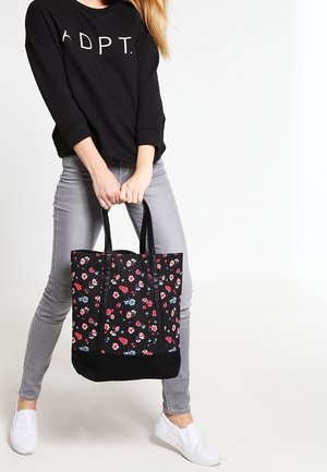 Svart blommig tote-väska med en blandning av röda och blå blommor, stadiga handtag och solid svart botten, kombinerad med grå jeans och vita slip-ons.